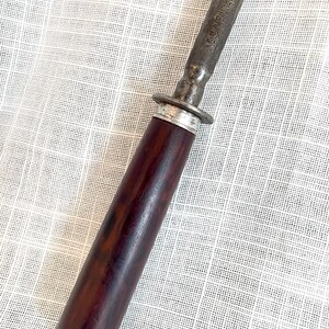May include: Close-up of a vintage steel knife sharpener with a dark brown wooden handle. The handle is cylindrical and has a silver metal ring near the blade. The blade is marked with the text "E.M. DICKINSON SHEFFIELD ENGLAND".