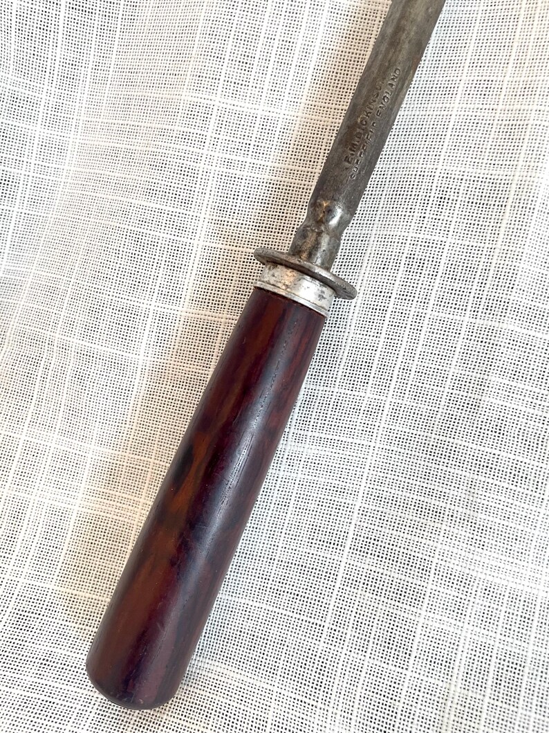 May include: Close-up of a vintage steel knife sharpener with a dark brown wooden handle. The handle is cylindrical and has a silver metal ring near the blade. The blade is marked with the text "E.M. DICKINSON SHEFFIELD ENGLAND".