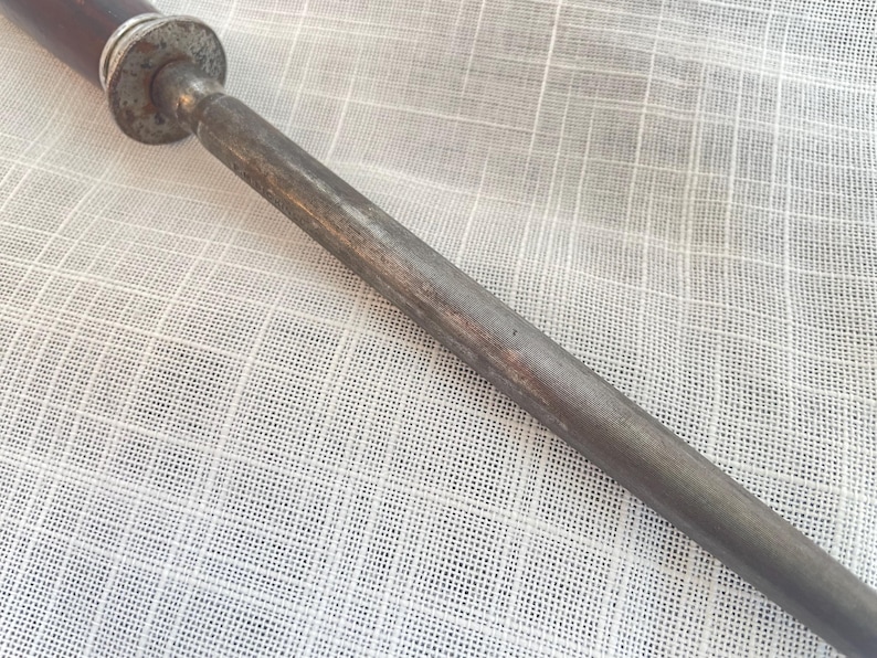 May include: Close-up of a steel knife sharpening tool with a dark brown handle and a textured, cylindrical metal rod. The tool has a worn, vintage appearance.