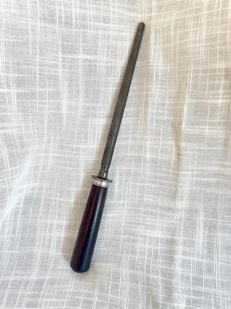 May include: A steel knife sharpening rod with a dark wooden handle and a silver-colored metal ring. The rod is tapered and appears well-used. The handle is a deep, rich brown color.