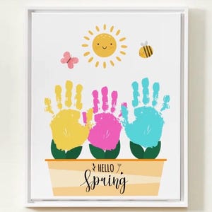 May include: A framed print featuring a spring-themed design. The artwork includes three colorful handprints in yellow, pink, and blue, placed above a planter. A smiling sun, a butterfly, and a bee are also depicted. The words "Hello Spring" are written in cursive.