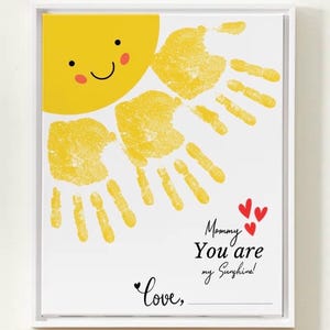 Könnte beinhalten: Gerahmter Druck mit einem gelben Sonnendesign, einem lächelnden Gesicht und Handabdruckstrahlen. Der Text lautet "Mama, du bist mein Sonnenschein!" mit roten Herzen und Platz für eine Unterschrift.