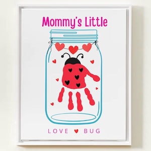 Op de afbeelding: Een ingelijste print met een potontwerp en een lieveheersbeestje van een handafdruk. De tekst "Mommy's Little" staat boven de pot en "LOVE BUG" eronder. Het lieveheersbeestje heeft een rode handafdrukbody met zwarte stippen en een zwarte kop.