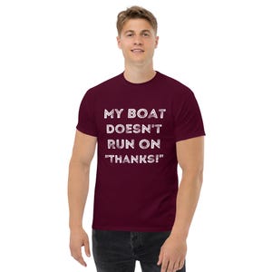 May include: A maroon t-shirt with the white text "MY BOAT DOESN'T RUN ON "THANKS!"" The shirt is a short-sleeved, crew neck style. The person in the image is wearing dark-colored pants.