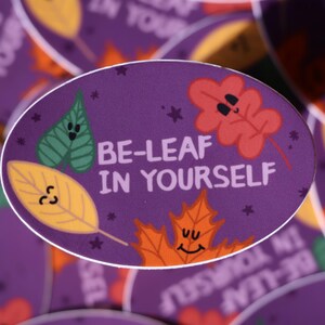 May include: Oval sticker with a purple background and the text "BE-LEAF IN YOURSELF". The sticker features cartoon leaves in green, yellow, and orange with smiling faces. Small star shapes are scattered around the leaves.