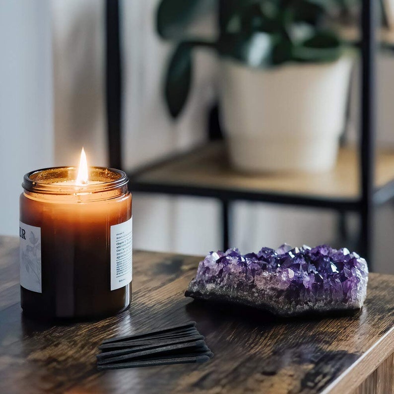 May include: A brown glass candle with a white label and a lit wick, a purple amethyst crystal cluster, and black matchsticks on a wooden table.