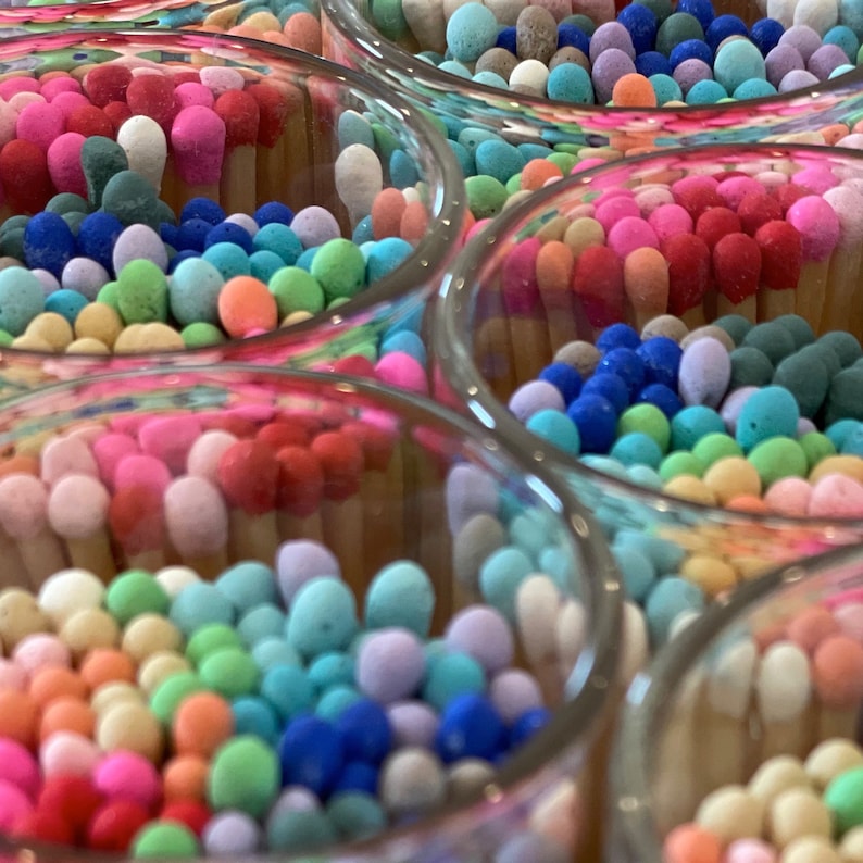 May include: Close-up of multiple glass jars filled with colorful matchsticks. The matchsticks are arranged in a circular pattern and have a variety of colors, including pink, blue, green, and yellow.