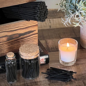 May include: Two glass jars filled with black matches, one with a cork lid and the other with a glass lid. A small pile of black matches is on the table next to the jars. A lit candle in a glass jar is on the table. A wooden box with black matches inside is in the background.