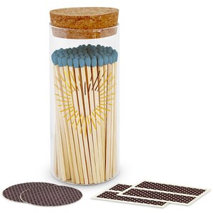 May include: A clear glass cylindrical jar filled with wooden matches featuring blue tips. The jar has a cork stopper and a yellow sunburst graphic printed on the front. Two circular dark brown striker pads and three rectangular striker strips are included.
