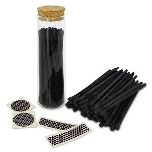 May include: A glass jar filled with black matches, with a cork lid. The matches are arranged in a bundle next to the jar, and there are four small, round, white stickers with a black honeycomb pattern.