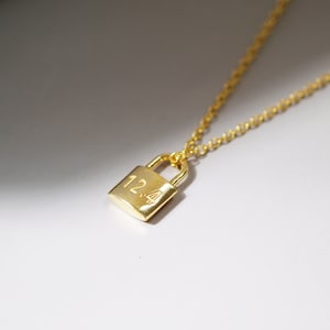 Może przedstawiać: Złoty naszyjnik z zawieszką w kształcie kłódki grawerowaną numerem "124".