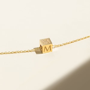 Op de afbeelding: Goudkleurige ketting met een kubusvormige hanger met de letter "M" gegraveerd op twee zijden. De hanger hangt aan een fijne ketting. De achtergrond is zacht en neutraal van kleur, met een schaduw over het oppervlak.