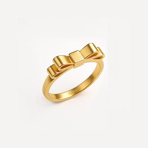 May include: A gold-colored ring featuring a bow design. The bow is centered on the ring's band, with the word "Ginny" engraved on the band. The ring is set against a white background.