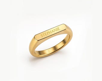 May include: A gold-colored ring with the name "STEPHANIE" engraved on the top. The ring has a rectangular top and a rounded band. The ring is set against a white background.