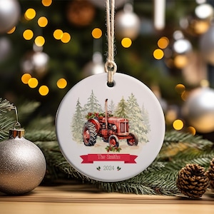 May include: A round, white ceramic Christmas ornament with a red tractor in a winter scene. The ornament has a red ribbon with the text "The Smiths 2024" printed on it.