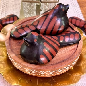 Puede incluir: Dos figuras de pájaros de cerámica con cuerpos negros y patrones de alas marrón rojizo descansan sobre una bandeja de madera. La bandeja se asienta sobre un plato decorativo dorado en forma de pétalo, creando una exhibición en capas.