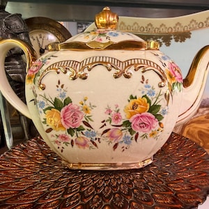 May include: Cream-colored porcelain teapot with a gold-trimmed lid and handle. The teapot is decorated with pink, yellow, and blue floral patterns. It sits on a decorative, brown, round plate.