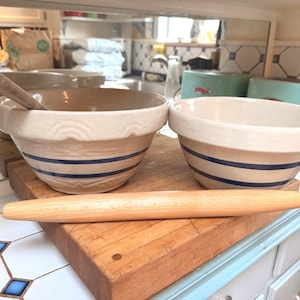 Puede incluir: Dos cuencos de cerámica con rayas azules y un rodillo de madera sobre una tabla de cortar de madera. Los cuencos son beige y blancos. Utensilios de cocina.