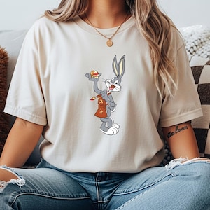 Peut inclure: T-shirt beige avec un dessin animé de Bugs Bunny. Le dessin animé montre Bugs Bunny tenant une pâtisserie et un pinceau avec un sac marron. Le t-shirt est de couleur neutre et a un col rond classique.