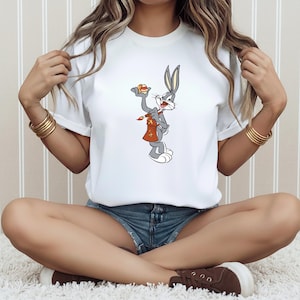 Peut inclure: T-shirt blanc avec une illustration de dessin animé de Bugs Bunny tenant une carotte et un pinceau. Le personnage de dessin animé porte un tablier rouge et tient une carotte. Le t-shirt est de style col rond classique.