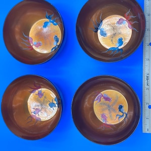 May include: Four decorative brown bowls with a gold-colored center. Each bowl features a hand-painted design of colorful fish. The fish are in shades of blue, red, and purple. The bowls are arranged in a square formation.