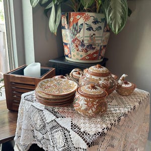May include: A ceramic tea set featuring a teapot, a lidded sugar bowl, and a stack of small plates, all decorated with intricate gold, orange, and brown dragon motifs on a cream background, displayed on a white lace tablecloth.
