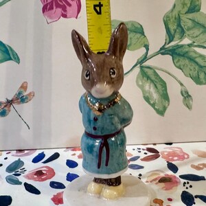 May include: A ceramic figurine of a brown rabbit wearing a teal coat with a gold necklace and a red belt. The rabbit is standing on a white circular base. The figurine is approximately 4 inches tall.