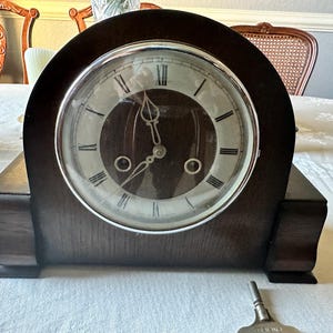 May include: A dark brown wooden mantel clock with a round face and Roman numerals. The clock has a silver-colored bezel and a key is resting in front of it. The clock is sitting on a white tablecloth.