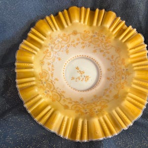 May include: A vintage, ruffled-edge bowl in a soft yellow hue. The bowl features a delicate floral pattern in a complementary shade of gold. The center of the bowl is a lighter, off-white color, with a small floral design. The bowl is likely made of glass.
