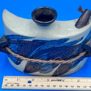 May include: A ceramic water vessel with a crescent shape, featuring blue and white glaze. The vessel has a dark brown spout and a rope handle. The design includes blue brushstrokes. The vessel is approximately 6 inches long.