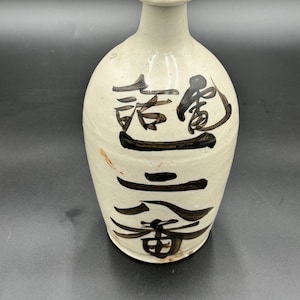 May include: A cream-colored ceramic bottle with a rounded body and a narrow neck. The bottle features black Japanese calligraphy. The bottle has a small opening at the top and appears to be handmade.