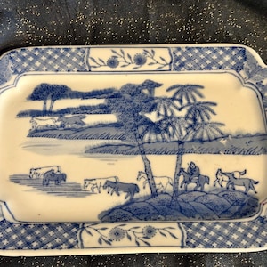May include: Rectangular ceramic tray with a blue and white pastoral scene. The tray features a detailed landscape with trees, figures on horseback, and animals. The border has a blue floral and geometric pattern.