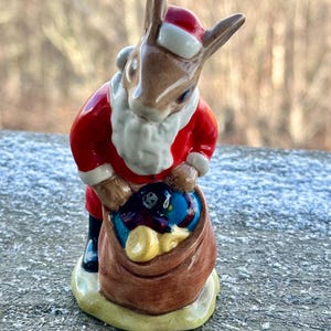 Puede incluir: Una figura de cerámica de un conejo vestido de Papá Noel, con un traje rojo con ribetes blancos y un gorro a juego. El conejo sostiene un saco marrón lleno de regalos coloridos. La figura está sobre una base verde.