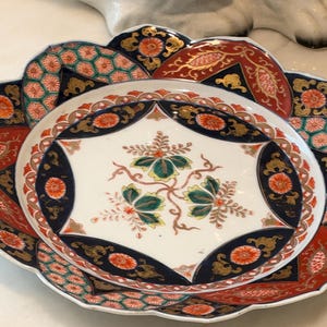May include: A decorative ceramic plate featuring a scalloped edge and a central white field with a green and orange floral motif. The border is adorned with intricate geometric patterns in navy blue, orange, and gold, with repeating circular flower accents.