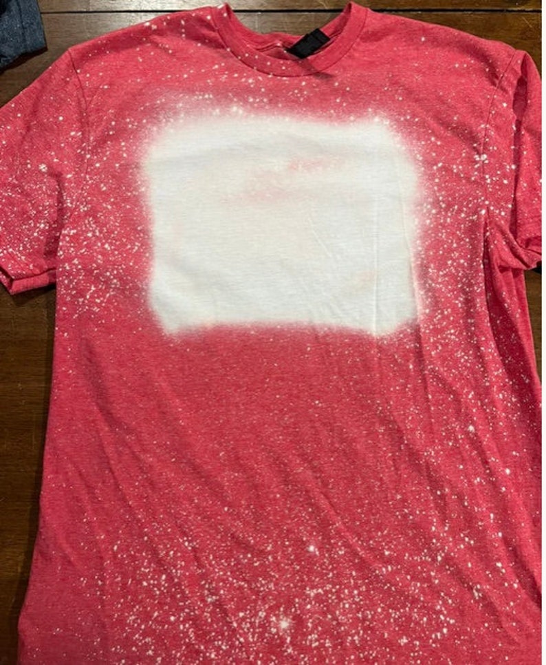 May include: Red t-shirt with a white bleach design. The design is a rectangle with a faded, speckled look.