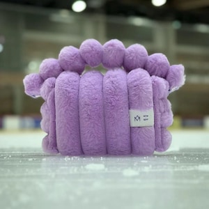 May include: A fluffy, lavender-colored handbag with a unique design. The bag features vertical, cylindrical sections and a top row of spherical accents. A small white tag with a logo is visible on the side. The bag is sitting on a light-colored surface.