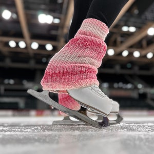 Puede incluir: Primer plano de patines de hielo blancos con cuchillas plateadas en una pista de hielo. Los patines se usan con calentadores de piernas de punto rosa y blanco. Se ven mallas negras encima de los calentadores.