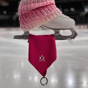 May include: A red fabric skate tassel with a silver ring and a sparkly 'M' design. The tassel is hanging from a pair of ice skates.