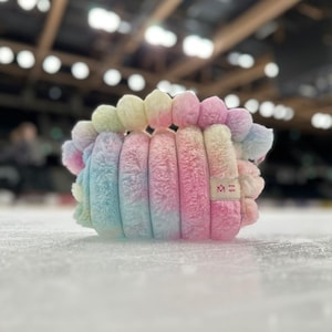 May include: A plush, rainbow-colored scrunchie with a soft, fuzzy texture. The colors blend from light blue to pink, yellow, and green. The scrunchie has a small tag with the words "wine spot" and a logo.