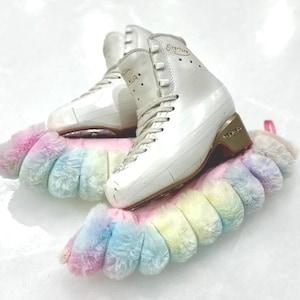 May include: White figure skates with gold accents and the word "Overture" on the side, resting on a pair of fluffy rainbow-colored skate guards.