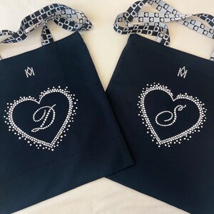 May include: Two black tote bags with white rhinestone heart designs, each with a different initial. The bags have patterned straps and a small initial above the heart. The background is a light beige.