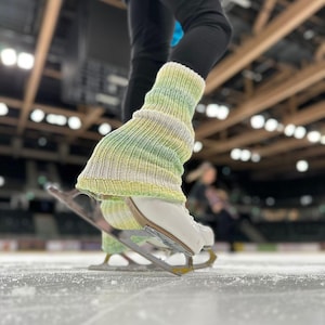 Op de afbeelding: Close-up van een been dat een paar witte en groene gebreide beenwarmers draagt met witte schaatsen. De persoon bevindt zich op een ijsbaan.