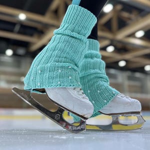 May include: A pair of teal blue knitted leg warmers with white beads, worn over white figure skates with silver blades.