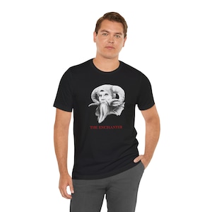 Peut inclure: T-shirt noir avec une illustration détaillée d'une figure avec de grandes cornes et une longue barbe. Le texte "THE ENCHANTER" est imprimé en rouge sous l'image. Le t-shirt est fait d'une matière douce.