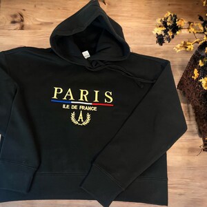 May include: Black hooded sweatshirt with the word "PARIS" embroidered in gold, with a blue and red stripe underneath. Below the word "PARIS" is the text "ILE DE FRANCE" and a gold wreath with the letter "A".
