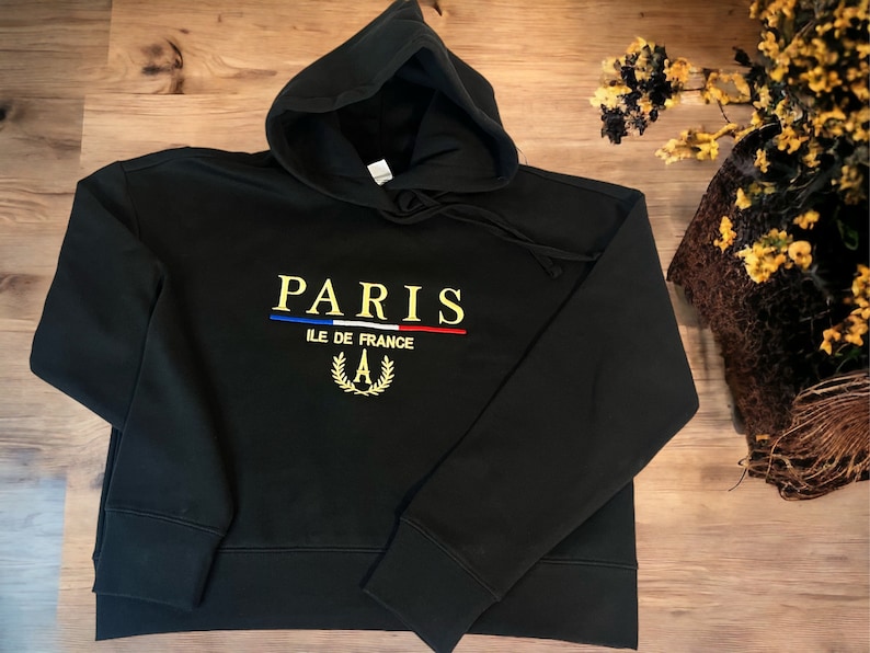 May include: Black hooded sweatshirt with the word "PARIS" embroidered in gold, with a blue and red stripe underneath. Below the word "PARIS" is the text "ILE DE FRANCE" and a gold wreath with the letter "A".