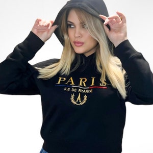 May include: Black hooded sweatshirt with gold embroidered "PARIS" text, a blue and red stripe, and a gold laurel wreath with the letter "A". The sweatshirt has long sleeves and a drawstring hood.