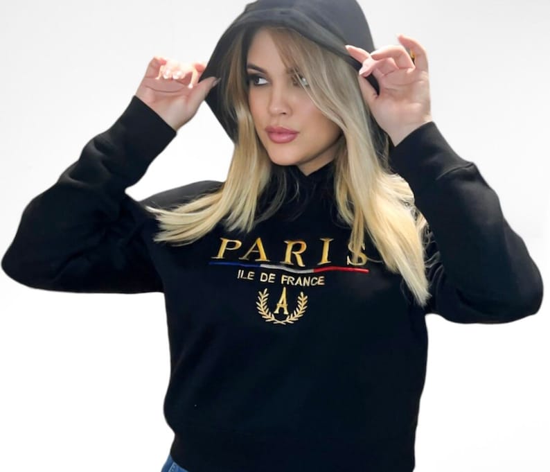 May include: Black hooded sweatshirt with gold embroidered "PARIS" text, a blue and red stripe, and a gold laurel wreath with the letter "A". The sweatshirt has long sleeves and a drawstring hood.
