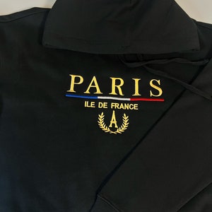 May include: Black hoodie with the word "PARIS" embroidered in gold thread. Below the word "PARIS" is a blue, white, and red stripe design. The hoodie also features the text "ILE DE FRANCE" and a gold wreath with the letter "A".