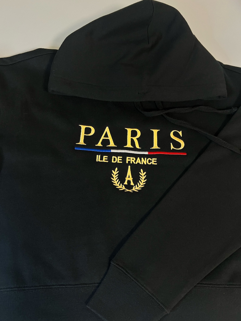 May include: Black hoodie with the word "PARIS" embroidered in gold thread. Below the word "PARIS" is a blue, white, and red stripe design. The hoodie also features the text "ILE DE FRANCE" and a gold wreath with the letter "A".